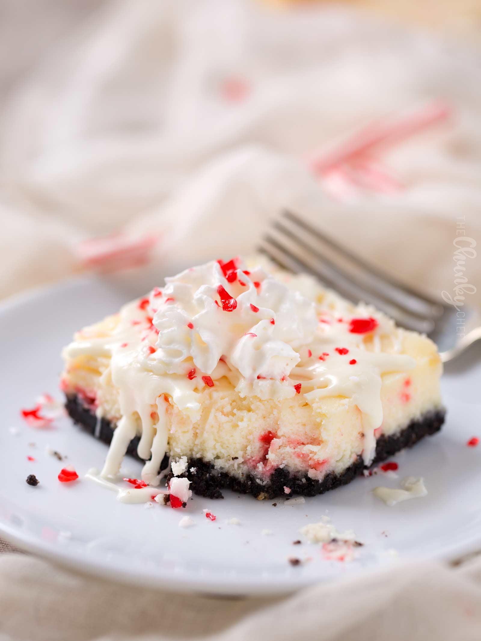 Peppermint White Chocolate Cheesecake Bars | An Oreo cookie crust, velvety smooth white chocolate peppermint cheesecake filling, candy cane pieces, and an extra drizzle of creamy white chocolate.  Holiday baking at it's finest! | The Chunky Chef | #cheesecakerecipes #cheesecakebars #peppermint #whitechocolate #holidaybaking #Christmasdesserts