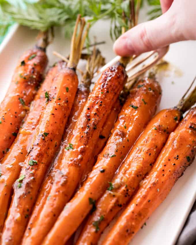 Tender baby carrots are coated in a sweet and savory glaze, then "roasted" in the crockpot! Free up your oven with this EASY side dish that's perfect for the holidays! | #carrots #sidedish #holidaysidedish #holidayrecipe #slowcookercarrots #crockpotcarrots using a fork to pick up glazed carrots from platter