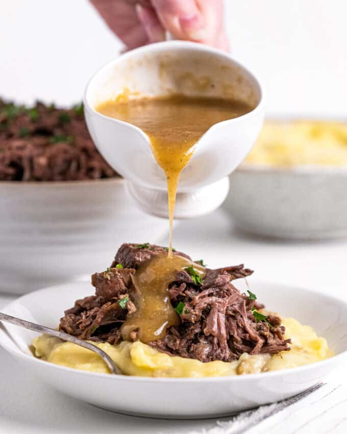 pouring gravy over pot roast meat on top of mashed potatoes