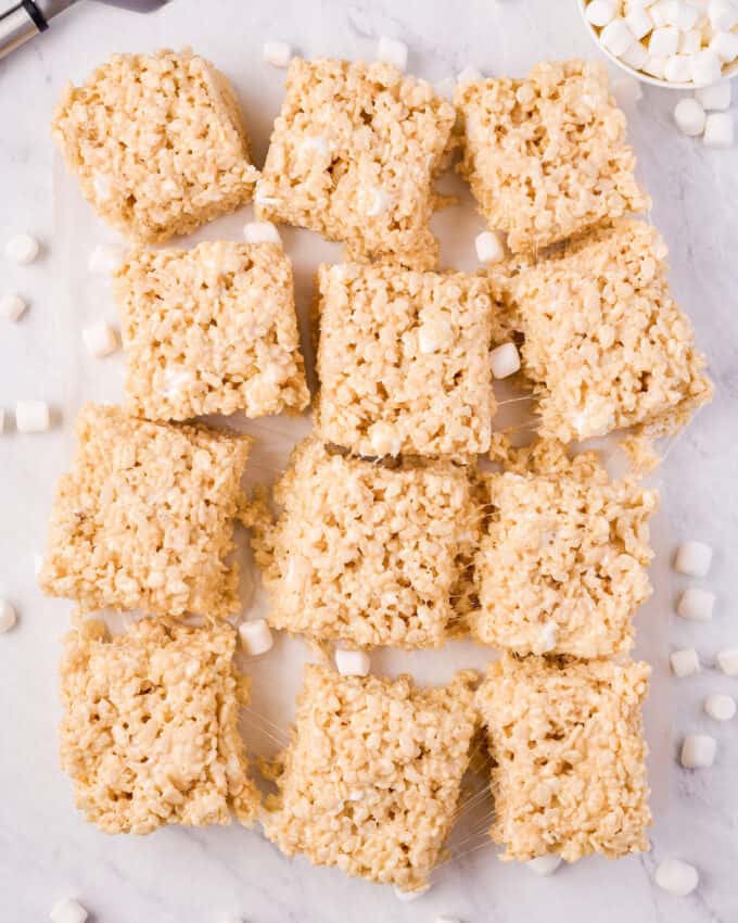 twelve krispie treat squares on marble counter with mini marshmallows scattered around