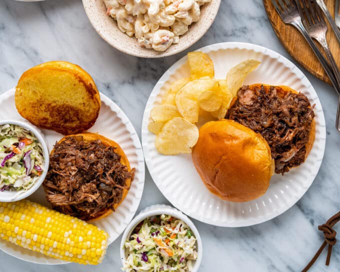 two plates with pulled pork sandwiches, potato chips, coleslaw, and corn on the cob