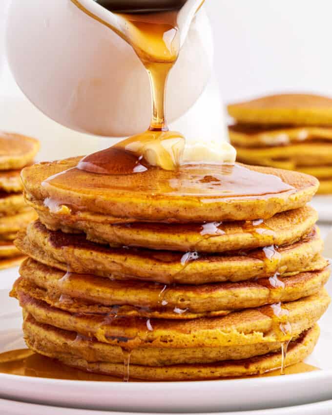 pouring maple syrup over a stack of pumpkin pancakes