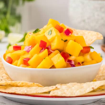 side view photo of a white bowl of mango salsa on a larger plate surrounded by tortilla chips