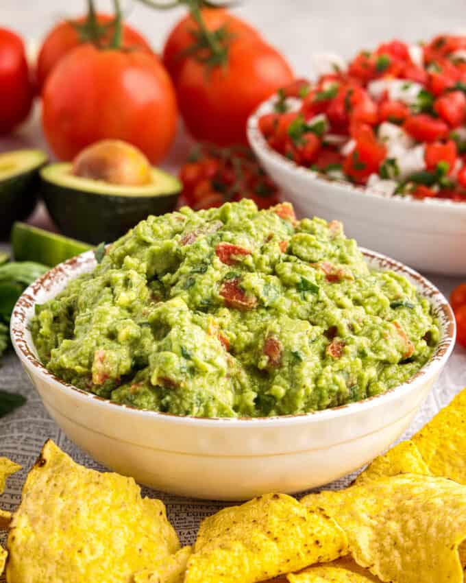 table with a bowl of guacamole dip surrounded by chips, avocados, limes, and tomatoes.