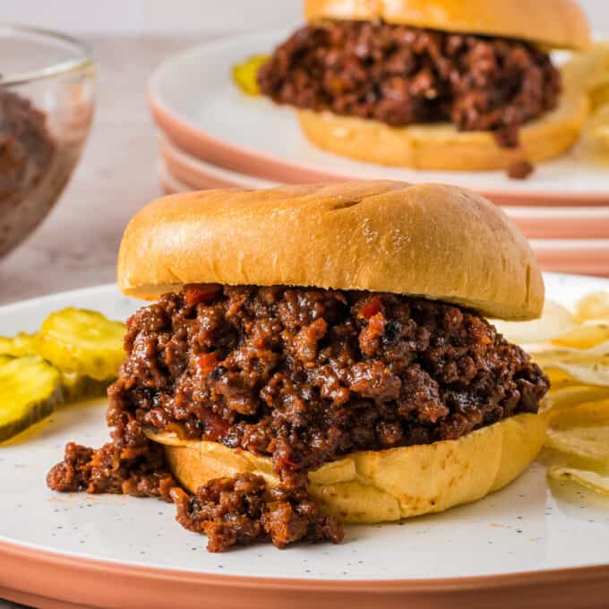 crockpot sloppy joes on buns on white plates with pickles and potato chips.