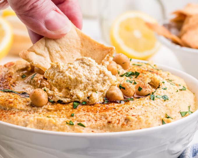 dipping a pita chip into white bowl of hummus.