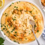 white bowl filled with hummus with chickpeas and olive oil, a spoon is scooping out some of it