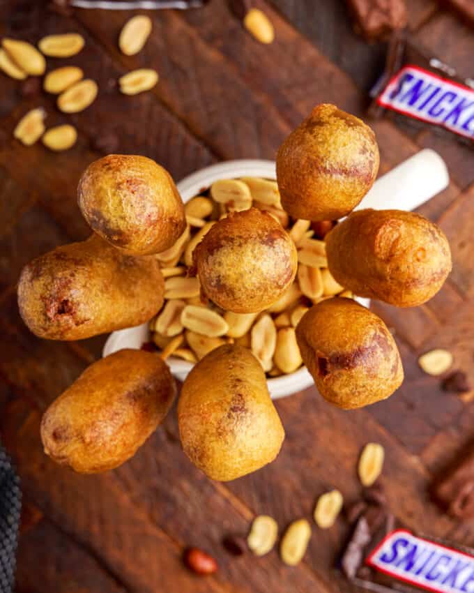 looking down on a wooden table with a jar filled with peanuts and fried snickers bars on sticks.