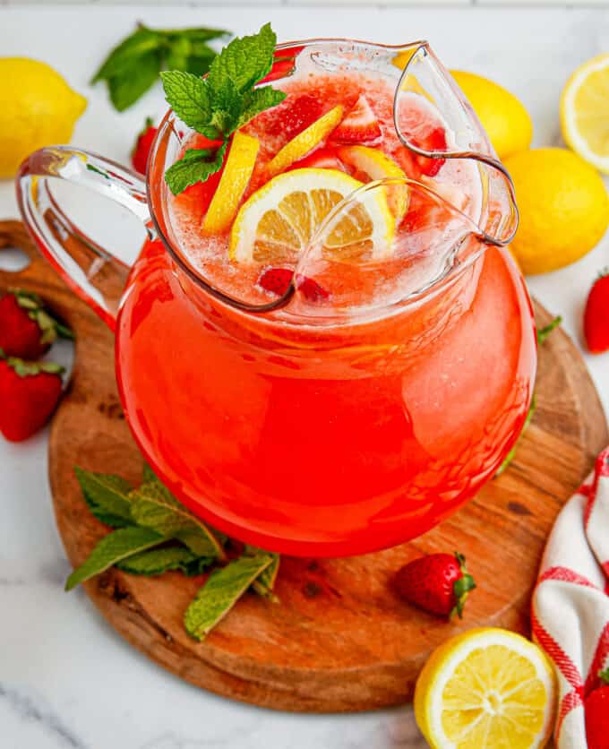 Glass pitcher of strawberry lemonade on a round wooden cutting board surrounded with lemons and strawberries.