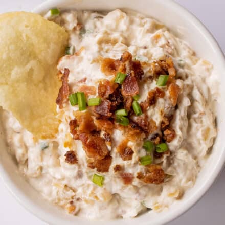 overhead photo of french onion dip in white bowl topped with bacon pieces and chives.