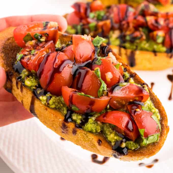 holding a slice of tomato bruschetta with pesto drizzled with balsamic glaze.