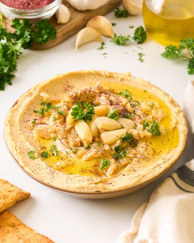 white table with bowl of garlic hummus surrounded by pita chips, garlic cloves, kitchen towel, etc.