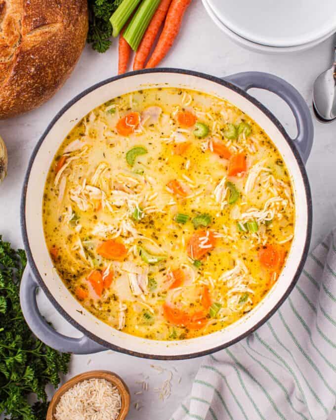 overhead photo of a dutch oven full of chicken and rice soup surrounded by a loaf of bread, linen napkins, and veggies.