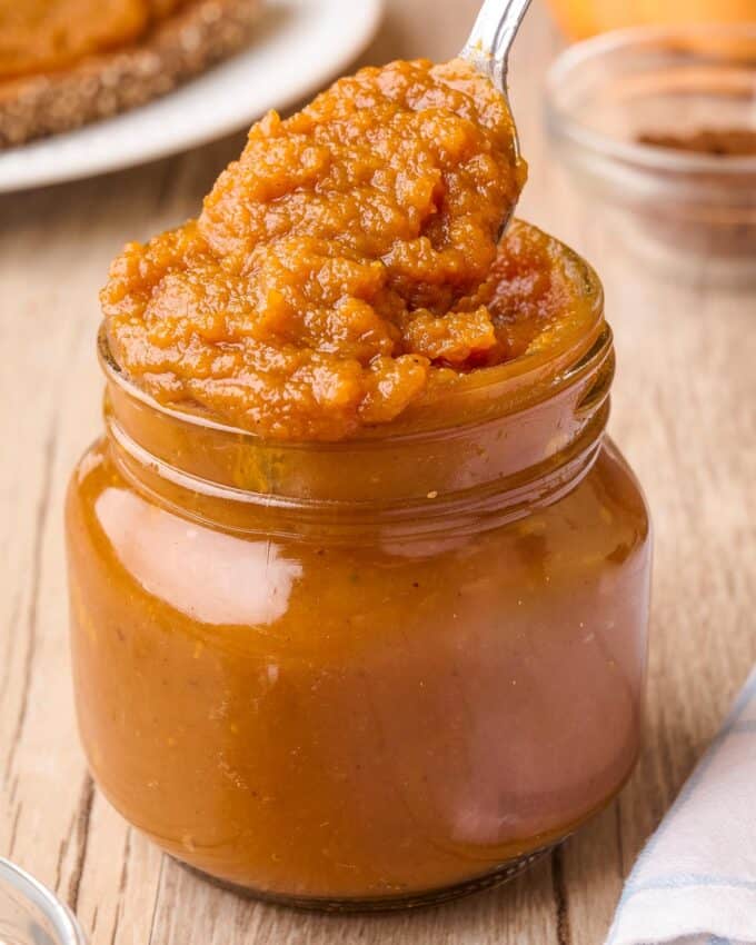 dipping a spoon into a glass jar filled with pumpkin butter.