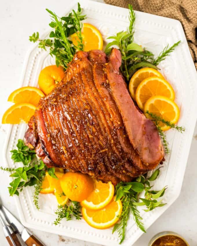 overhead view of a spiral sliced ham on a white platter with orange slices and fresh herbs.