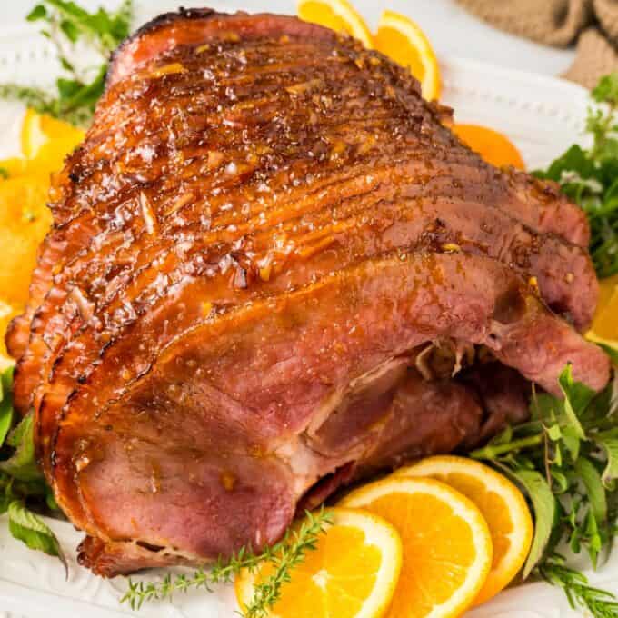 spiral ham with orange glaze on a white platter surrounded by orange slices and fresh herbs.