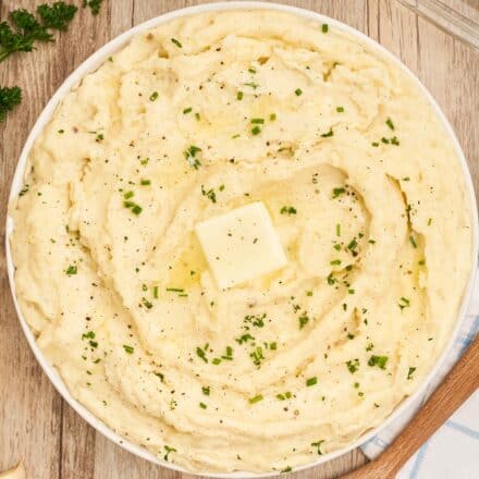 overhead photo of mashed potatoes in a white serving bowl topped with a pat of butter and fresh herbs.