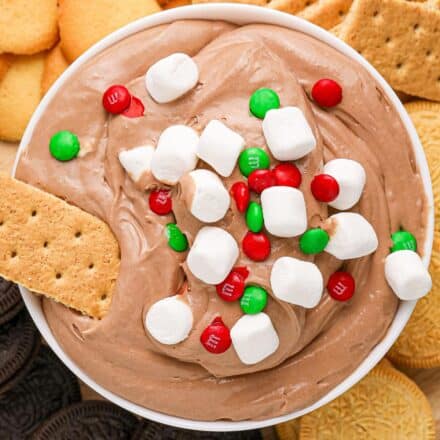 overhead photo of a white bowl of hot cocoa dip topped with marshmallows and m&m's, with a graham cracker dipped in it.