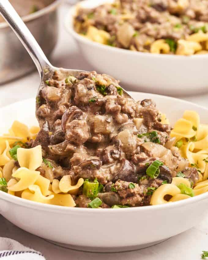 spooning beef stroganoff over egg noodles in a white bowl.