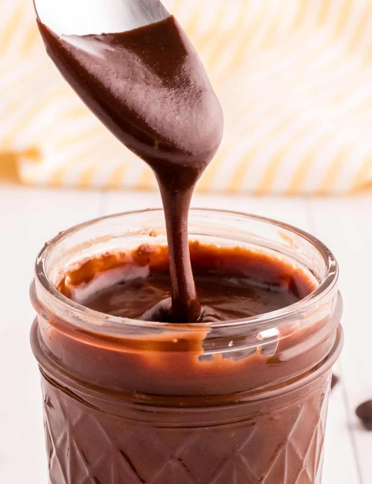 Glass jar of thick homemade hot fudge sauce with a spoon lifting a glossy chocolate drizzle on a white counter