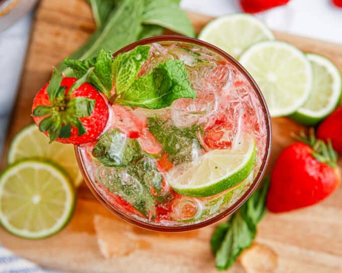 overhead view of a glass of mojito with strawberries on a wooden cutting board.
