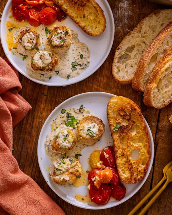 two white plates with creamy pesto scallops, toasted bread, and burst cherry tomatoes.