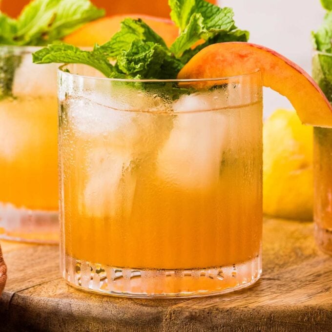 rocks glass of peach bourbon smash on a wooden board garnished with mint and peaches