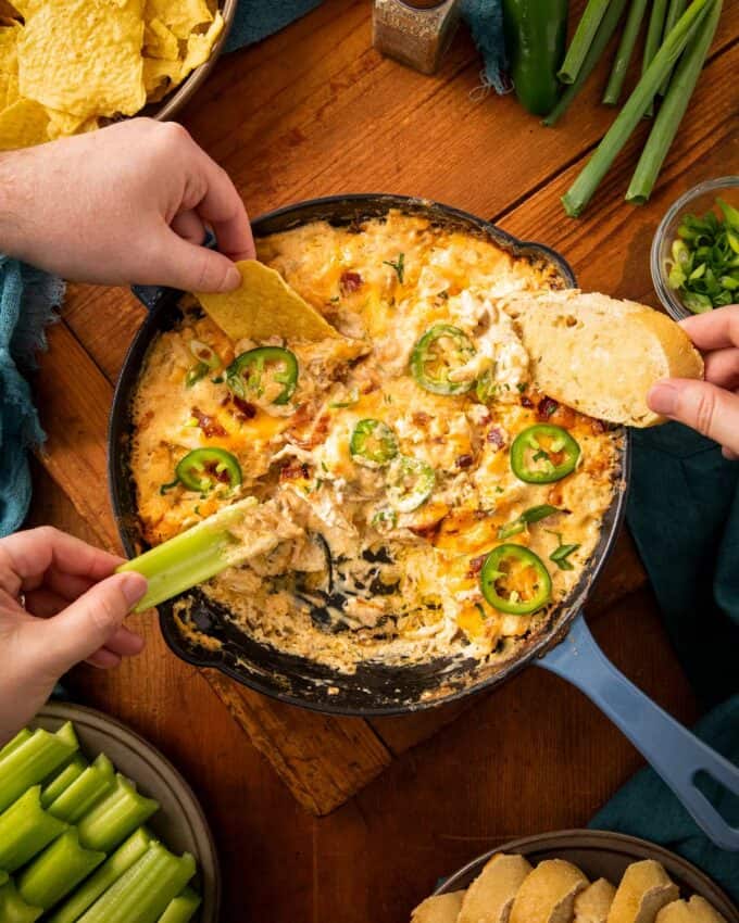 hands dipping chips, celery, and bread into chicken jalapeno popper dip at a party