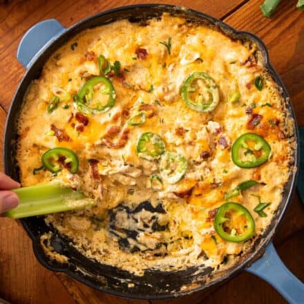 dipping celery stalk into chicken jalapeno popper dip in a cast iron skillet